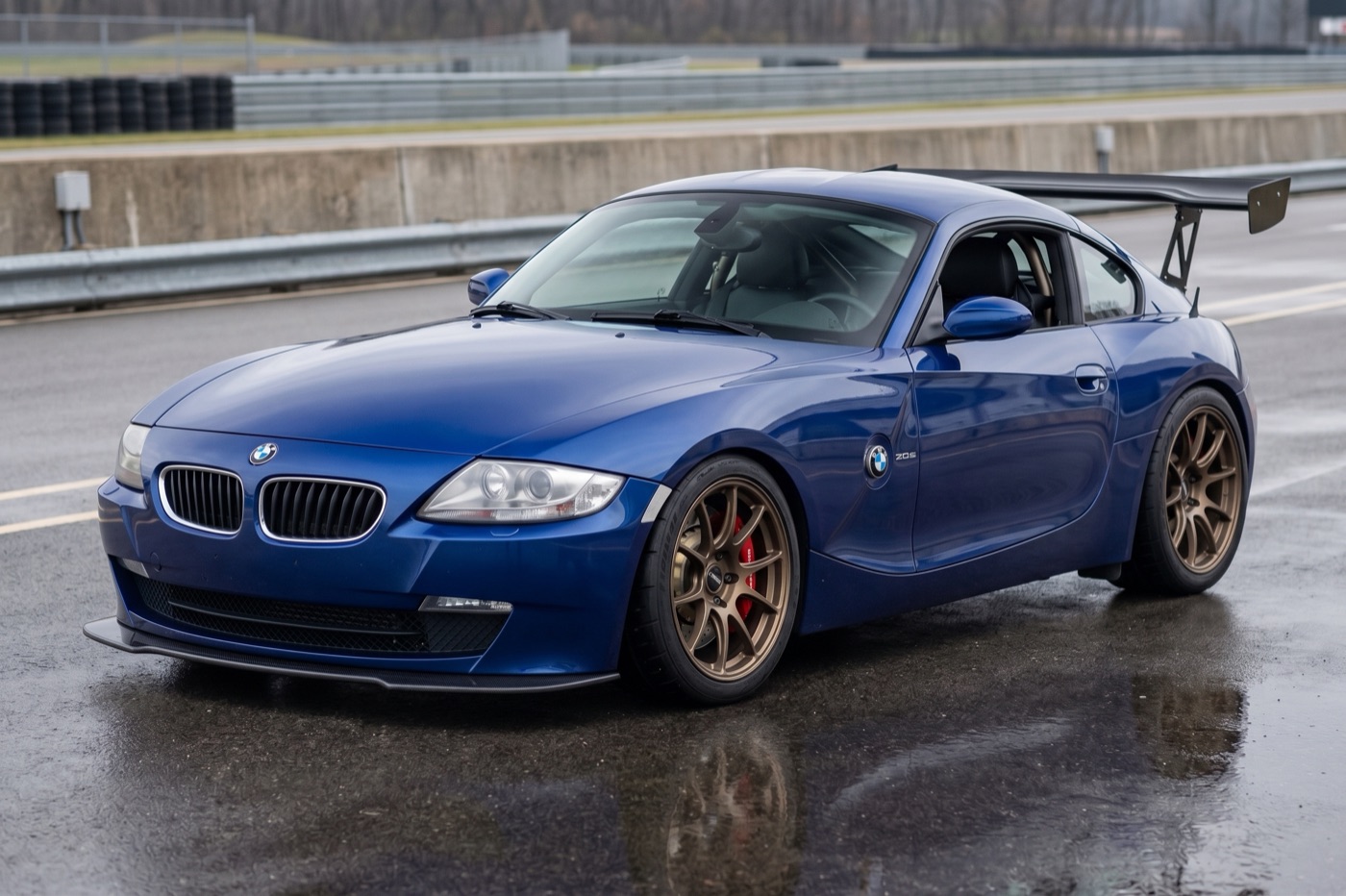 AI-generated render of the 2008 Z4 Coupe as a finished track car: Montego Blue, bronze forged wheels, red big-brake calipers, carbon front splitter and a tall rear wing, on a wet pit lane